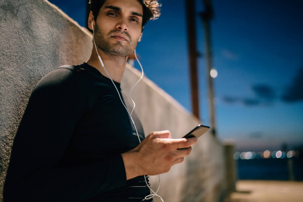 Cropped portrait of male jogger in earphones choosing audio from music playlist standing outdoors in evening time.Sportsman in black active wear for workout installing mobile player - Foto, Bild