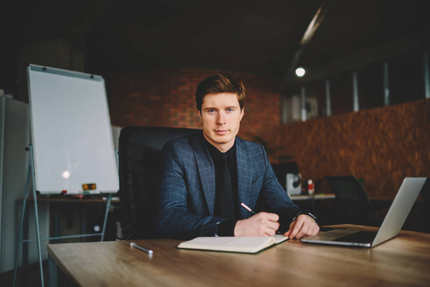 Half length portrait of proud ceo in formal suit making notes of corporate statement sitting at desktop in cabinet,confident owner of company creating business plan in netbook looking at camera - Фото, зображення