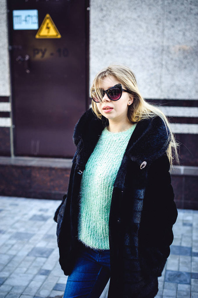 young woman in a fur coat, a sweater and fashion sunglasses with ears, walking around the city. Street fashion - Foto, Imagen
