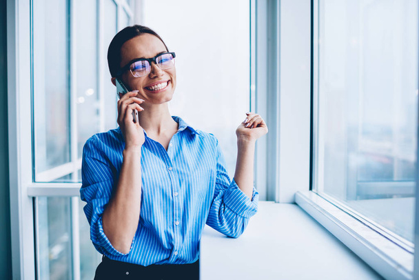 Cheerful businesswoman in spectacles satisfied with good news talking on mobile phone with partner, happy overjoyed female in formal wear smiling during smartphone conversation about winning tender - Photo, Image