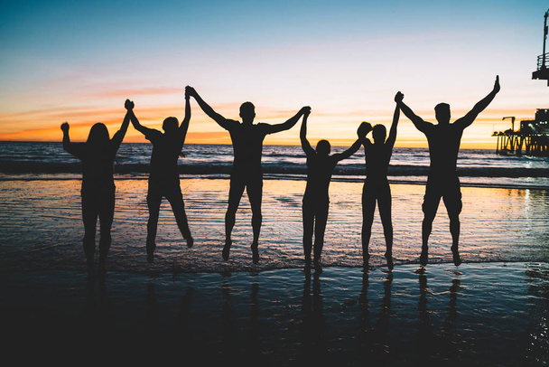 Szczęśliwy młodych osób płci męskiej i żeńskiej trzymając się nawzajem ręce stojący przy plaży w krajobraz zachód słońca, sylwetka grupy osób korzystających lato wakacje razem i skoki w powietrzu, pojęcie przyjaźni - Zdjęcie, obraz