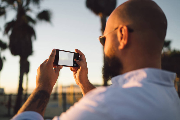 Vista posterior de un blogger masculino usando un teléfono inteligente con pantalla simulada para tomar fotos y compartir en redes, un tipo hipster sosteniendo un teléfono móvil moderno haciendo fotos durante las vacaciones de verano por la noche
 - Foto, imagen
