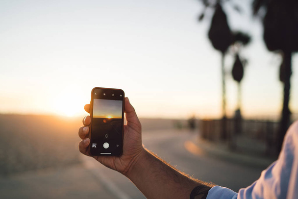 Close up view of male shooting video of sunset and skyline on smartphone camera, cropped image of man's hand holding mobile phone using app for making pictures of sundown during summer vacation - Foto, imagen