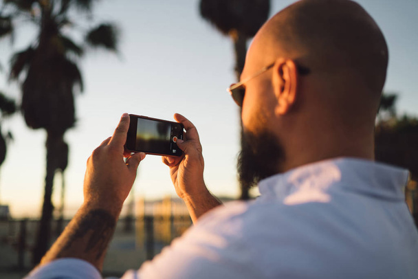 Close up rear view of young male holding in hands smartphone with modern camera for taking photos of beautiful sunset, cropped view of hipster guy shooting evening landscape on mobile telephone - Photo, image