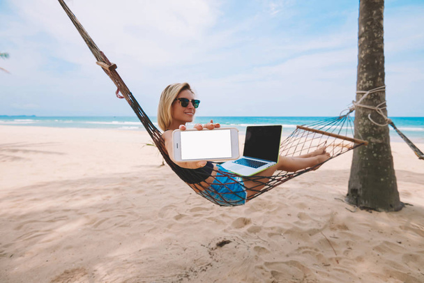 Freelancer turístico feminino mostrando smartphone com tela em branco para aplicação ou site de emprego empreendedor enquanto trabalhava remotamente no computador portátil moderno durante as férias de verão na praia
 - Foto, Imagem