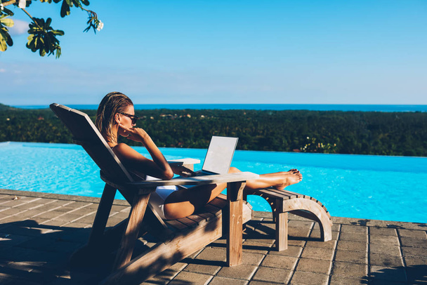 Pensive slim young woman working freelance at netbook with blank screen area while resting on sunbed near swimming blue poor enjoying summer resort.Tourist reading news on laptop while sunbathing - Photo, Image