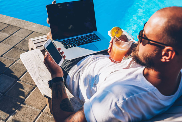 Young businessman with cocktail in hand reading notification on smartphoe while resting on sunbed near swimming pool and doing remote job at modern laptop computer with mock up area with your content - Fotoğraf, Görsel