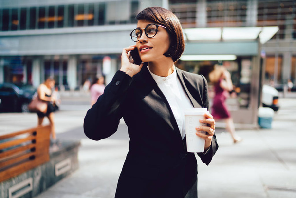 Confident businesswoman in trendy eyewear having mobile conversation standing on street with tasty beverage in hand.Serious female trader talking with service operator strolling with in new york city - Φωτογραφία, εικόνα