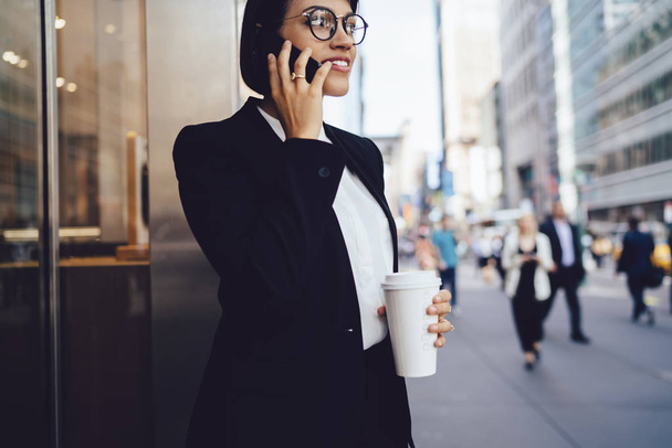 Cropped image of prosperous professional lawyer in eyeglasses answering on incoming phone call strolling with tasty coffee in metropolis of modern city.Cheerful female banker talking on cellular - Foto, Bild