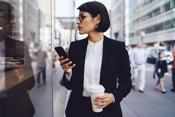 Serious female trader in eyewear walking on street of megalopolis with tasty coffee in hands.Confident businesswoman in stylish formal wear looking away holding mobile telephone and coffee to go - Foto, immagini
