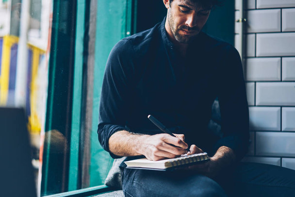 Cropped image of stylish casual dressed young man writing down text information in notepad sitting in coffee shop interior.Pensive student in black shirt noting checklist in notebook - Фото, зображення