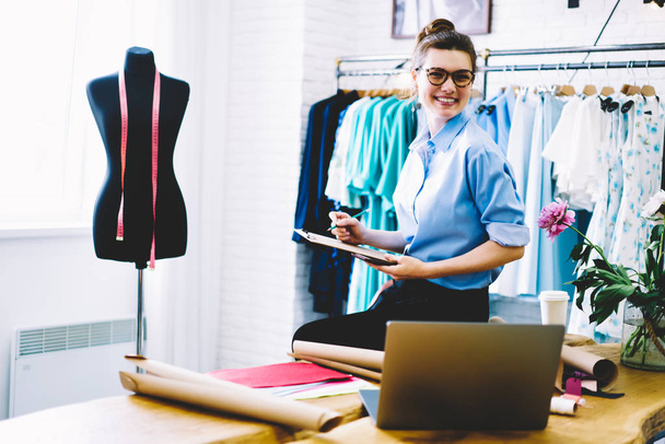 Smiling female designer in eyeglasses looking at camera while making notes of measurements working in showroom on collection of stylish clothing sitting on desktop with garment accessories and netbook - 写真・画像