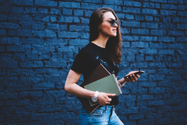 Beautiful female blogger strolling at urban setting and chatting with followers in social network on modern telephone while standing against wall with promotional background for your advertising - Photo, Image