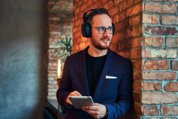 Portrait of a handsome bearded male wearing stylish suit listening music with the tablet against a brick wall. - Fotó, kép