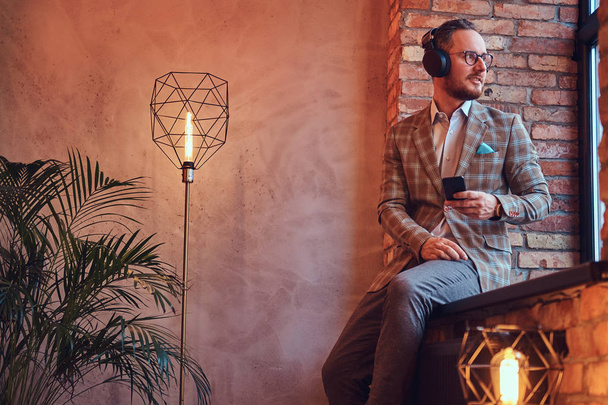 Retrato de um homem elegante em um terno de flanela e óculos ouvindo música com o telefone e fones de ouvido sentados em uma soleira da janela em um quarto com loft interior
. - Foto, Imagem
