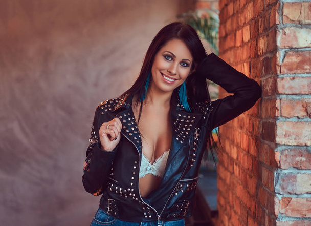 Portrait of a sexy sensual brunette posing in stylish leather jacket and jeans leaning against a brick wall. - Fotografie, Obrázek