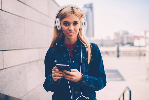 Half length portrait of attractive blonde student looking at camera while listening audio lesson of learning foreign languages in modern headphones connected to smartphone device standing outdoors - Valokuva, kuva