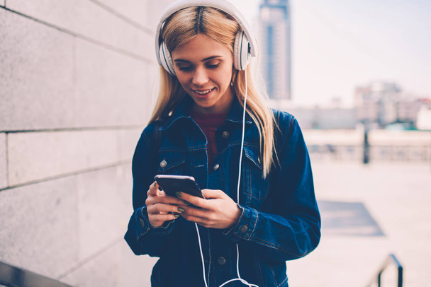 Smiling hipster girl in denim jacket reading incoming notification on smartphone while listening audio songs in modern headphones walking in urban setting.Cheerful student chatting online on telephone - 写真・画像