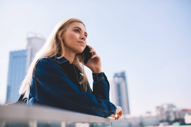 Beautiful female traveller talking with operator on telephone while standing outdoors on bridge.Attractive young woman with blonde hair communicaing on modern smarthone during walk in metropolis - 写真・画像