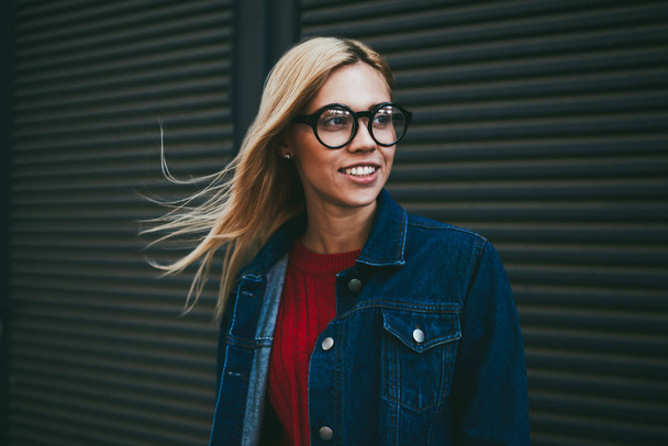 Positive young woman with blonde hair laughing while looking away outdoors.Hipster girl in optical spectacles dressed in stylish denim apparel standing on black promotional background - Φωτογραφία, εικόνα