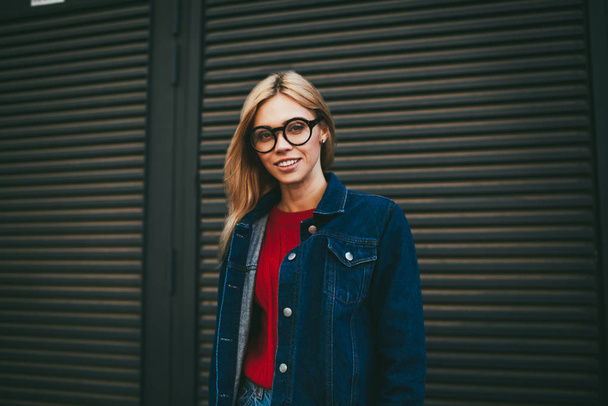 Half length portrait of happy stylish young woman with blonde hair smiling at camera while posing on promotional background with copy space.Cheerful student in optical eyeglasses and denim clothes - Foto, immagini