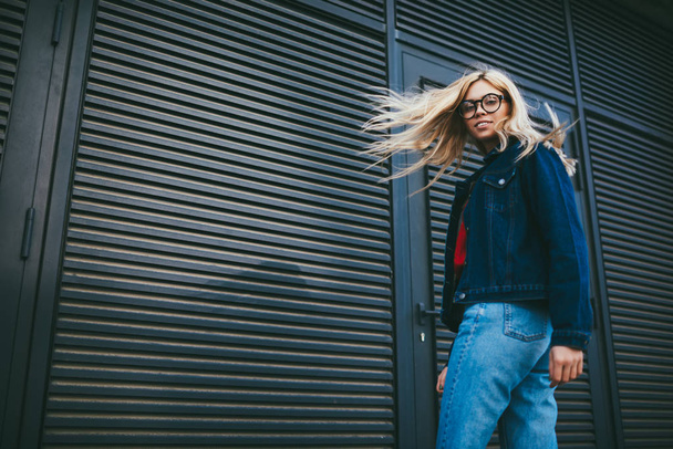 Portrait of cheerful stylish woman with gorgeous blonde hair smiling at camera while standing on promotional background.Positive hipster girl in eyewear dressed in denim apparel near copy space area - Foto, Imagem