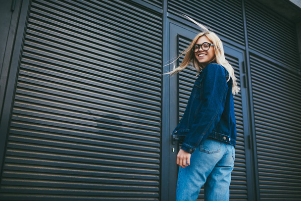 Half length portrait of happy blonde young woman in stylish eyeglasses smiling at camera.Cheerful hipster girl dressed in denim apparel while standing on promotional backgrounf with publicity area - Photo, Image