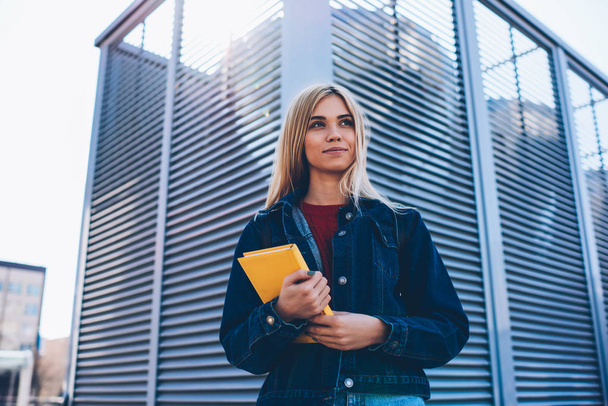 Young gorgeous positive female student dressed in trendy denim clothing recreating outdoors while holding yellow notepad.Cute pondering hipster girl looking away during spending free time at street - Fotografie, Obrázek