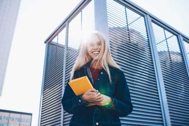 Young attractive blonde female student enjoying recreation at spring sunny urban setting during strolling.Beautiful prosperous stylish hipster girl spending free time holding yellow notepad outdoors - Foto, imagen