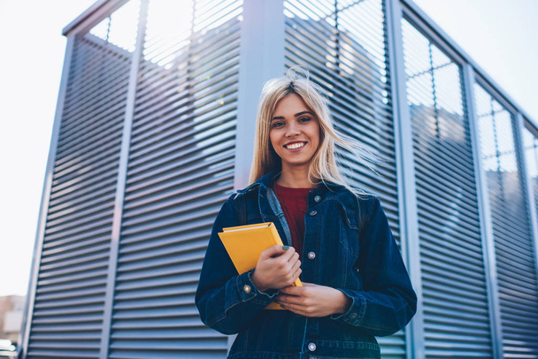 Half length portrait of attractive smart cheerful student enjoying free time at sunny urban setting during walk.Cute successful blonde woman looking at camera while waiting for meeting outdoors - 写真・画像