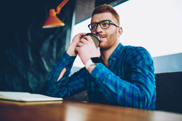 Handsome young man with red hair drinking tasty coffee to go resting in coworking space.Hipster guy in optical eyeglasses and casual styish shirt enjoying beverage sitting in cafeteria interior - Foto, Imagen