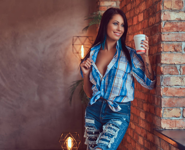 A sexy brunette in a flannel shirt and jeans holds a cup of coffee posing against a brick wall.  - Фото, зображення