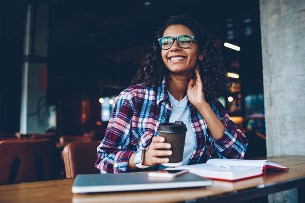 Dreamy smiling african american student thinking on literature plot of book while holding tasty coffee to go in hand during break.Positive dark skinned teen with curly hair pondering on creative ideas - Foto, Imagen