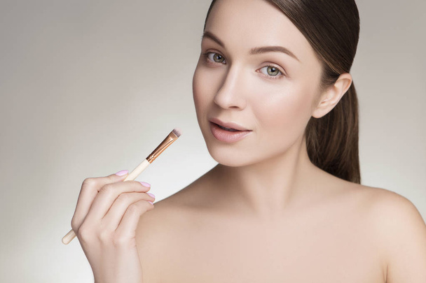Beautiful brunette caucasian young woman prepare herself, applying lipstick on her lips with a brush. Clean, fresh, natural, flawless skin. Soft smile on her face. Close up on a neutral background - Photo, image