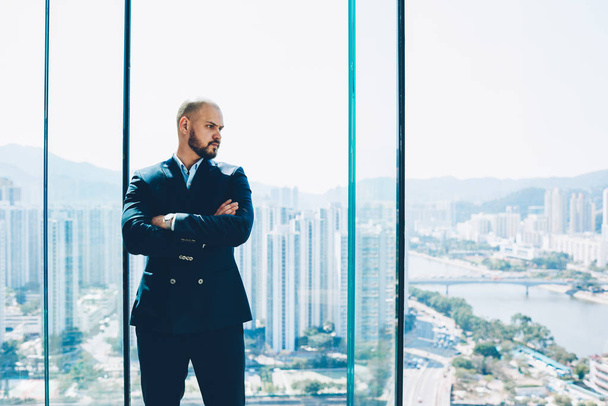 selbstbewusster bärtiger Geschäftsmann in schwarzer Festkleidung und mit Blick aus dem Fenster mit Großstadt im Hintergrund. - Foto, Bild