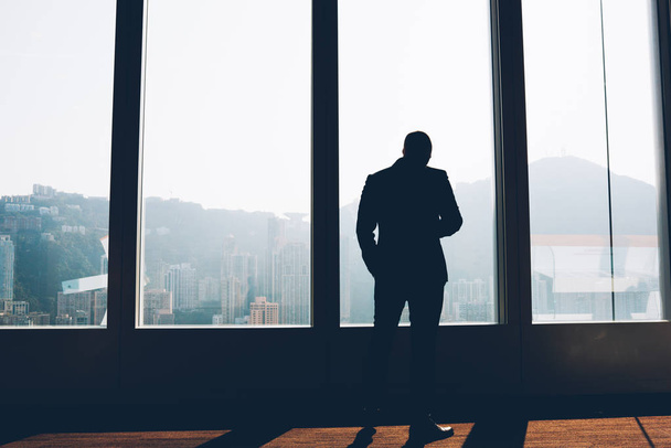 Back view of businessman in elegant suit standing indoors in loft interior.Male entrepreneur dressed in formal wear looking out of window enjoying scenery and thinking on building project in office - Foto, imagen