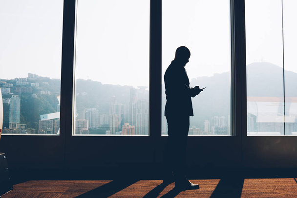 Black silhouette of financial manager standing near big window and downloading file with accounting reports on mobile phone.Male proud ceo reading last business news on smartphone standing in office - Foto, Imagen