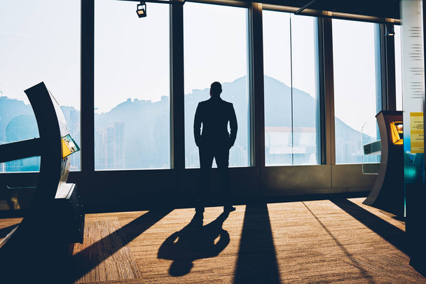 Black view of male entrepreneur dressed in formal apparel looking out of glass window with amazing scenery and thinking on solution of business project enjoing sunset after hard day - Photo, Image