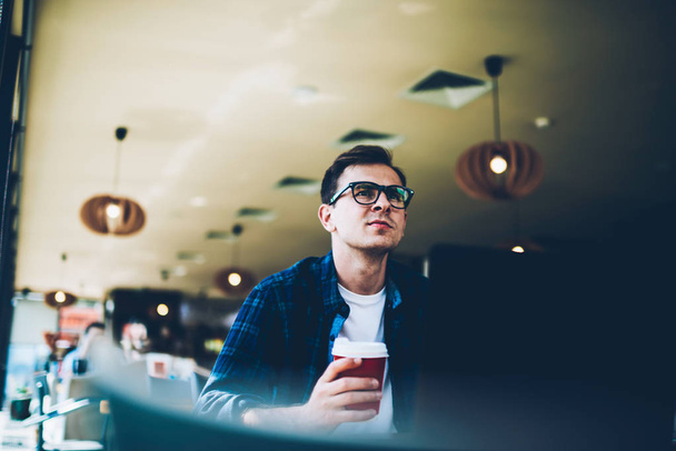 Good looking talented student in trendy eyewear enjoying delicious coffee during break in preparation for coursework.Handsome hipster guy holding take away beverage while sitting at table indoors - Foto, Bild