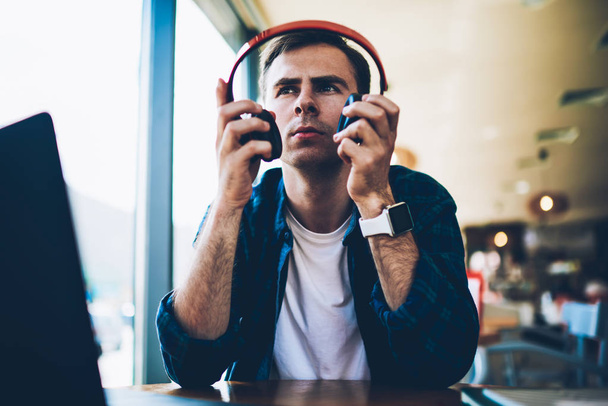 Young handsome pondering male student taking off big modern headphones while recreating sitting at wooden table indoors.Good looking man putting on head device for listening favorite music in cafe - Фото, изображение