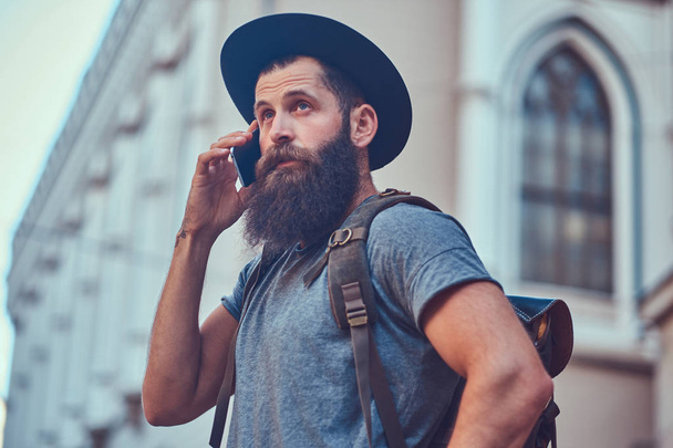Komea hipster-matkustaja, jolla on tyylikäs parta ja tatuointi käsivarsillaan pukeutuneena rentoihin vaatteisiin, seisoo kadulla puhumassa puhelimessa.
. - Valokuva, kuva
