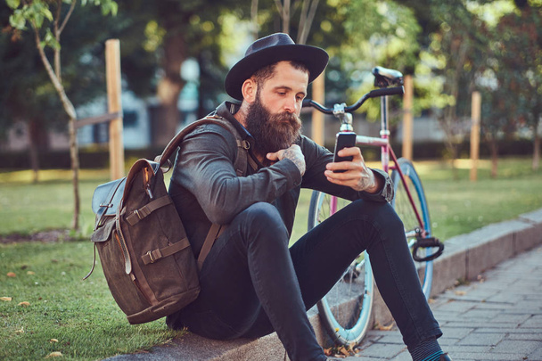 Guapo viajero hipster con una elegante barba y tatuaje en sus brazos vestido con ropa casual y sombrero con una bolsa, utilizando el teléfono inteligente, sentado en la acera, descansando después de un paseo en bicicleta
. - Foto, imagen