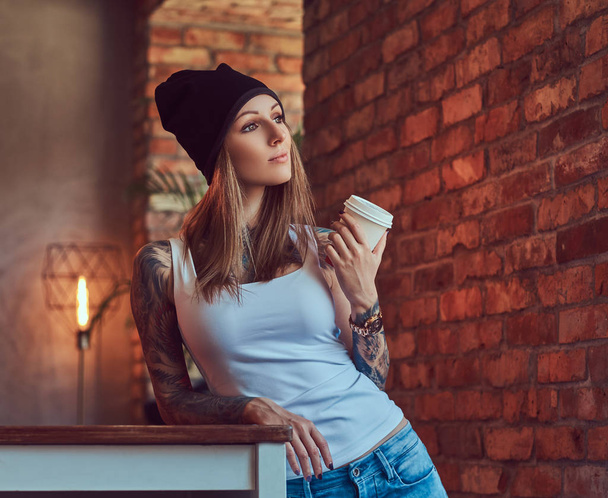 Een getatoeëerde sexy blonde in een t-shirt en een hoed met kopje koffie in een kamer met loft interieur. - Foto, afbeelding