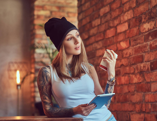 Uma loira sexy tatuada em uma camiseta e um chapéu com xícara de café e tablet em um quarto com interior loft
. - Foto, Imagem