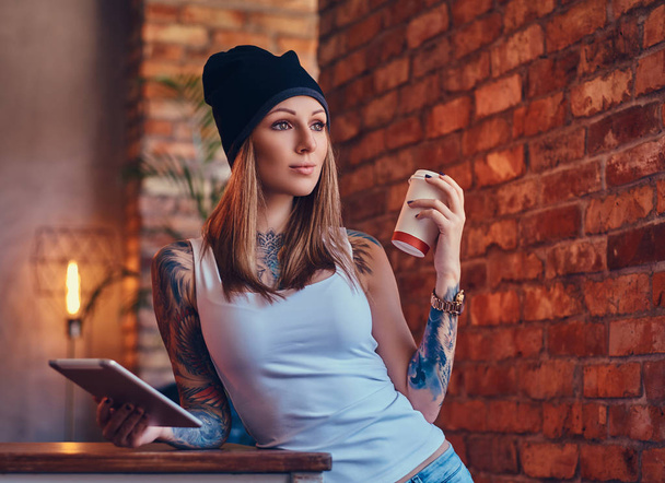 A tattooed sexy blonde in a t-shirt and a hat with cup of coffee and tablet computer in a room with loft interior. - Photo, Image