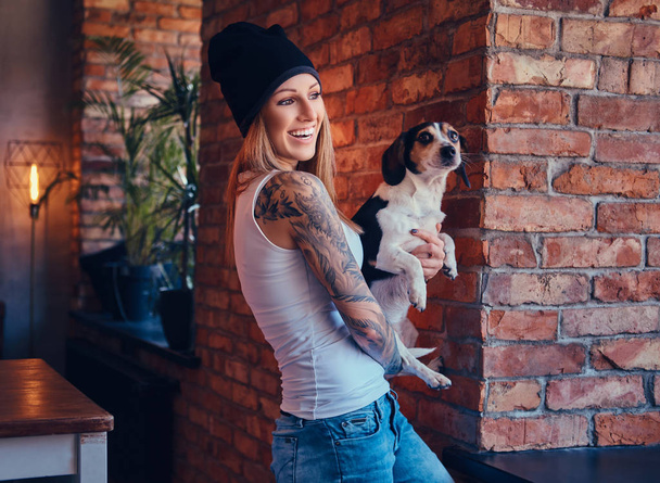 Стильна татуйована блондинка в футболці і джинсах тримає милий собаку
. - Фото, зображення