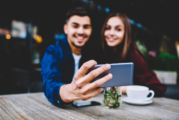 Selective focus on smartphone in hand of young man sitting with girlfriend on cafe terrace posing for selfie, cheerful male and female hipsters making picture for share in networks using mobile phone - Foto, Imagen