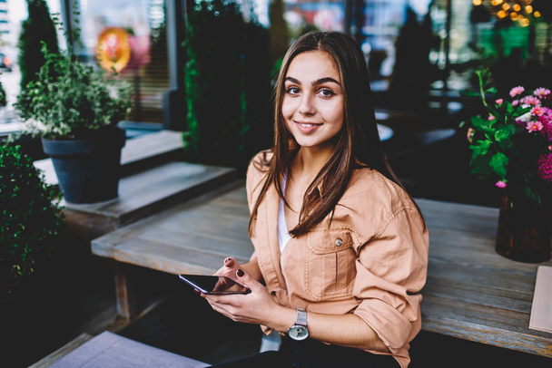 Portrait of cute female student resting in cafe interior sending message via smartphone, pretty young hipster girl in casual wear looking at camera while using popular application on mobile phone - Foto, imagen