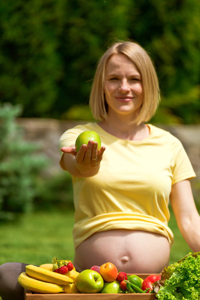 Pregnant woman sitting on grass and holding out apple in hand - Φωτογραφία, εικόνα