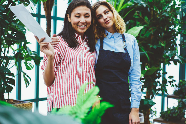 Portrait of prosperous colleagues working in orangery in friendly atmosphere, woman proprietor of glasshouse planning budget for business consulting with female assistant in apron looking at camera - Foto, Bild